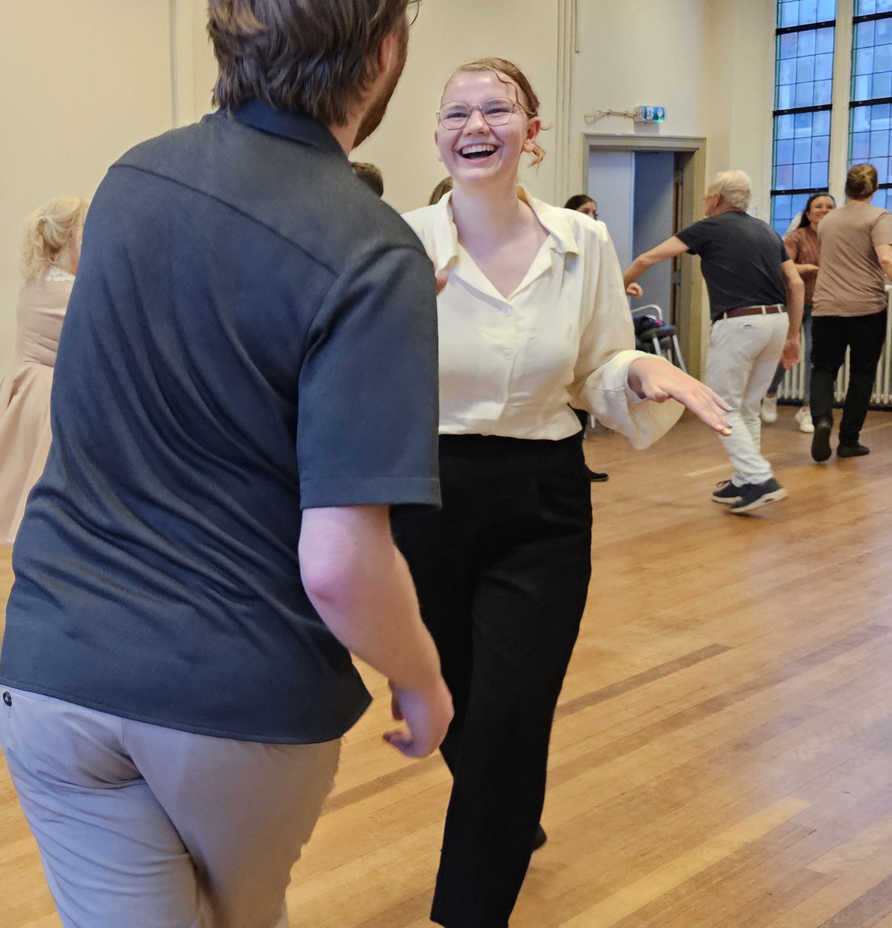 Jeffrey en Renée lachen en dansen samen in een vrolijke lesruimte terwijl ze Lindy Hop voordoen aan een groep dansers. Ze zijn het nieuwe docentenduo bij JazzOut Den Bosch.